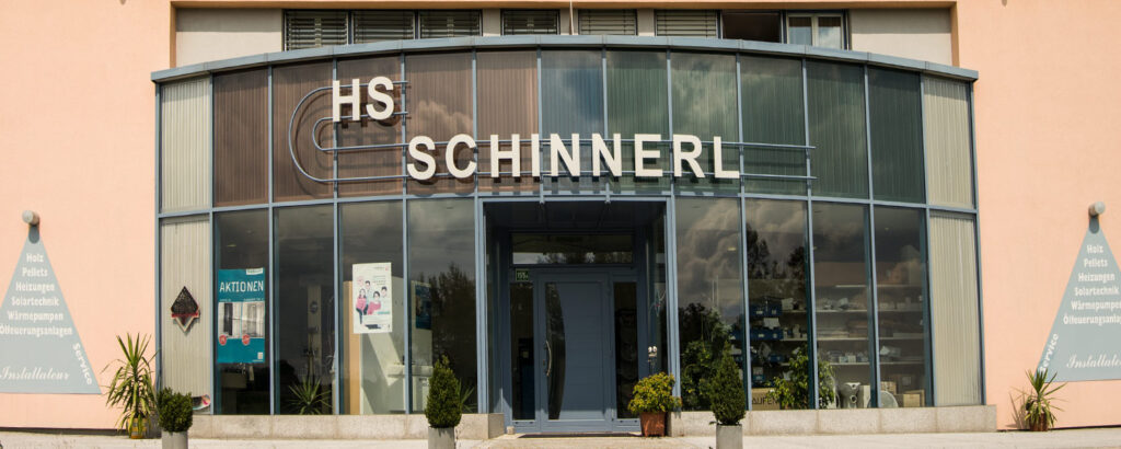 Foto des zweistöckigen Firmengebäudes. Blaue Glasfassade mit weißer Aufschrift: "HS Schinnerl".