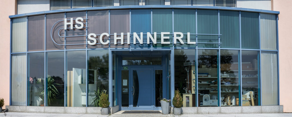 Foto des zweistöckigen Firmengebäudes. Blaue Glasfassade mit weißer Aufschrift: "HS Schinnerl".